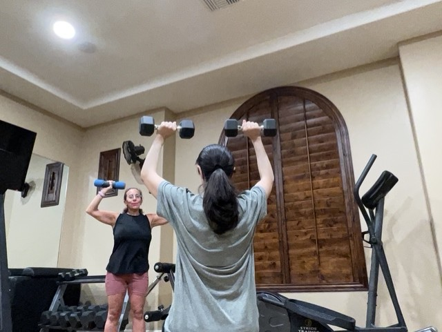 Linda coaching her client Fatima through a dumbbell overhead press in a home gym