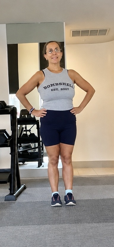 Linda in 2026 standing strong in a fitness studio wearing a Bombshell tank top and navy shorts